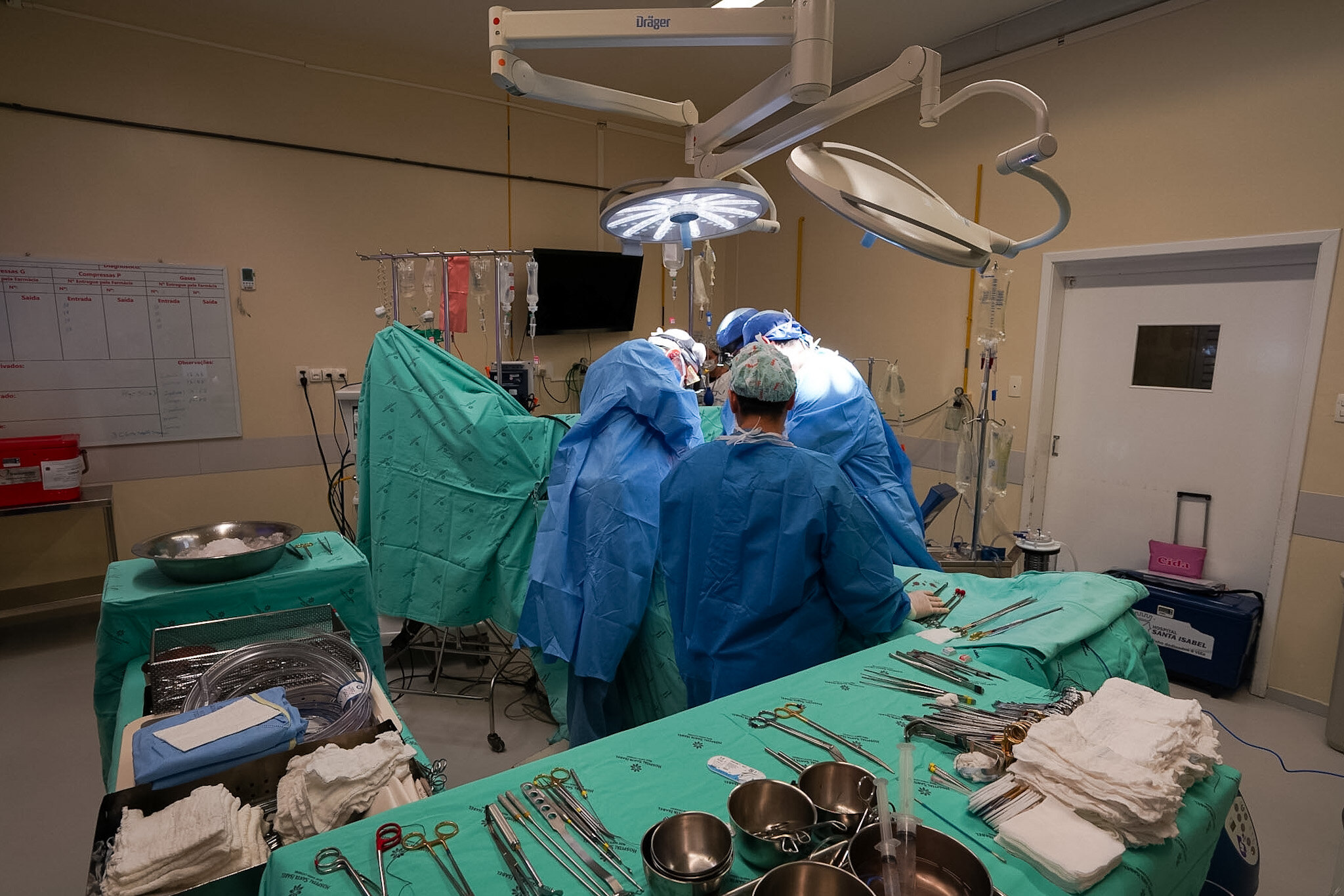 Equipe médica realiza procedimento cirúrgico em uma sala de cirurgia totalmente equipada. Quatro profissionais vestindo aventais e toucas estéreis concentram-se sobre o paciente, coberto por campos cirúrgicos verdes. A sala está iluminada com luzes cirúrgicas pendentes e há diversos instrumentos cirúrgicos organizados sobre uma mesa ao primeiro plano, incluindo pinças, tesouras, gazes e bandejas metálicas. Ao fundo, observam-se bolsas de soro penduradas, equipamentos médicos, uma lousa branca e uma caixa térmica vermelha, típica para transporte de órgãos. O ambiente é limpo, controlado e altamente técnico.