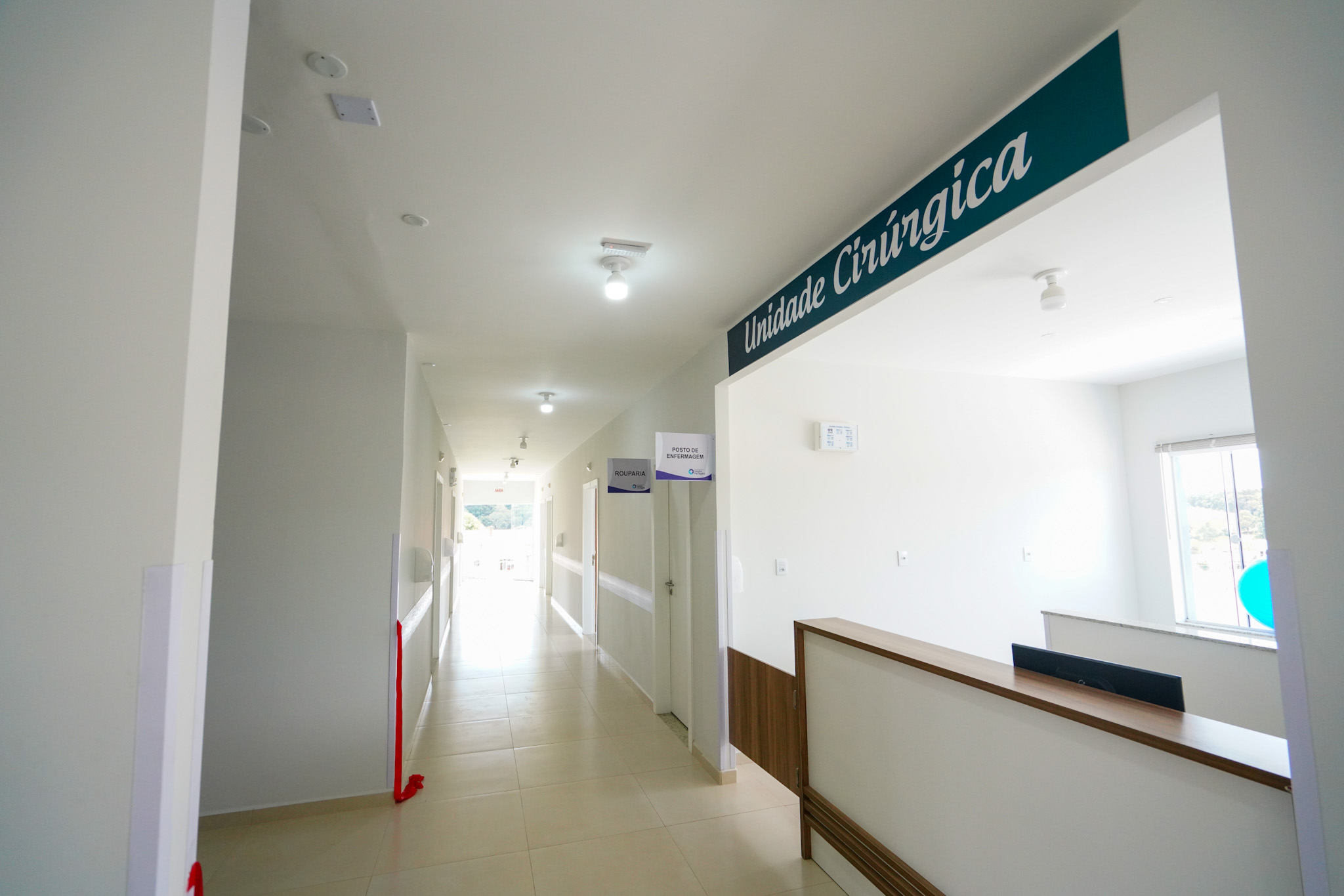Corredor interno de uma unidade de saúde com piso claro e paredes brancas. À direita, há um balcão de recepção em madeira e vidro fosco, abaixo de uma placa com a inscrição "Unidade Cirúrgica". Mais adiante no corredor, há placas identificando setores como "Rouparia" e "Posto de Enfermagem". A iluminação é feita por lâmpadas embutidas no teto e, ao fundo, uma janela permite a entrada de luz natural. Ambiente limpo e organizado, típico de uma instalação hospitalar.
