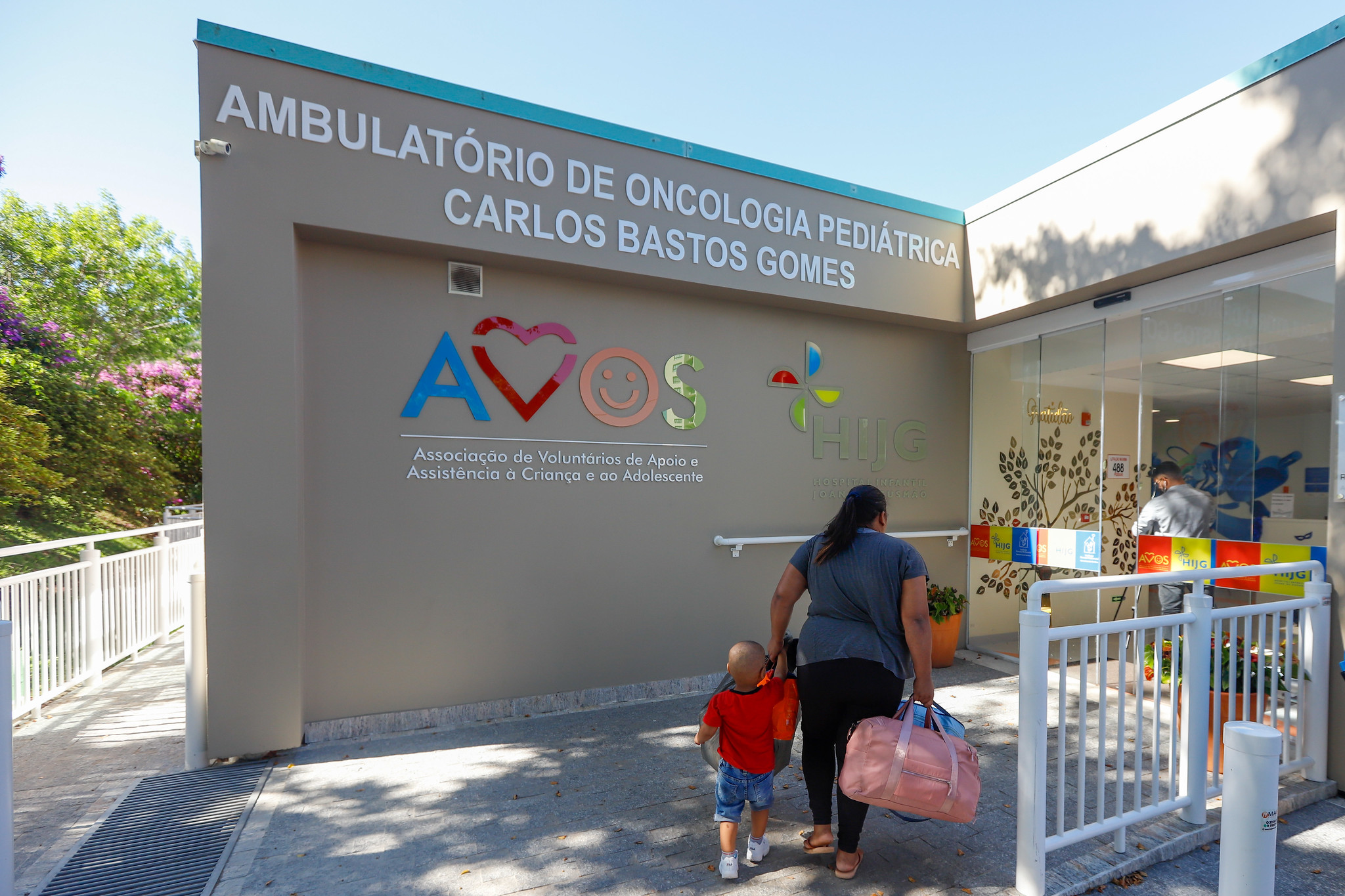 Fachada do Ambulatório de Oncologia Pediátrica Carlos Bastos Gomes, com logos da AVOS e do Hospital Infantil Joana de Gusmão. Uma mulher segura a mão de uma criança pequena careca e carrega bolsas ao entrar no prédio, sugerindo um momento de acompanhamento para tratamento médico infantil.