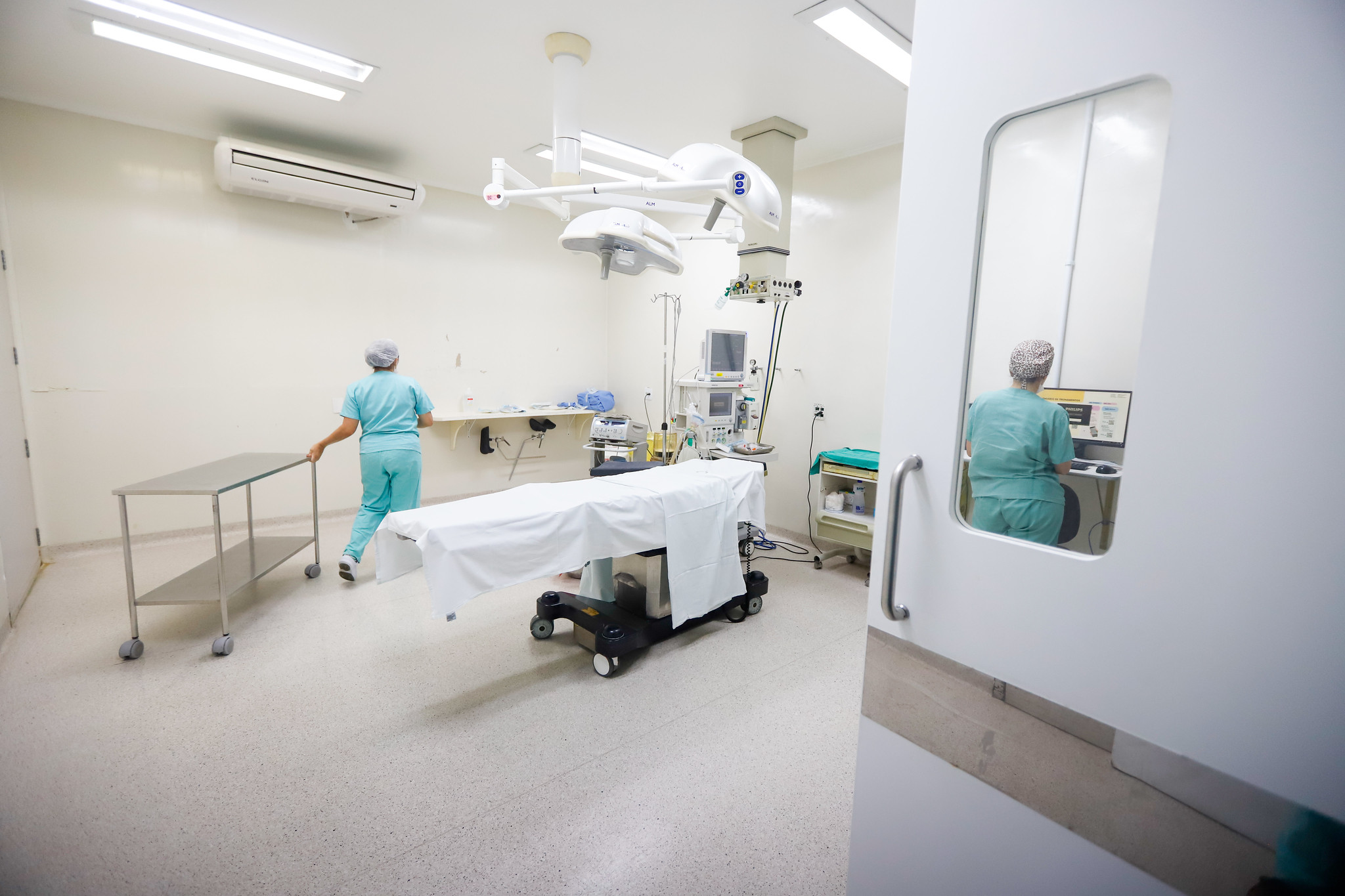 Ambiente de centro cirúrgico limpo e bem iluminado. Ao centro, há uma maca coberta com lençol branco, rodeada por equipamentos hospitalares. À esquerda, uma profissional da saúde empurra uma mesa de apoio de inox. À direita, outra profissional sentada de costas trabalha em um computador, visível por uma porta com visor de vidro. O ambiente transmite organização e preparo para procedimentos médicos.