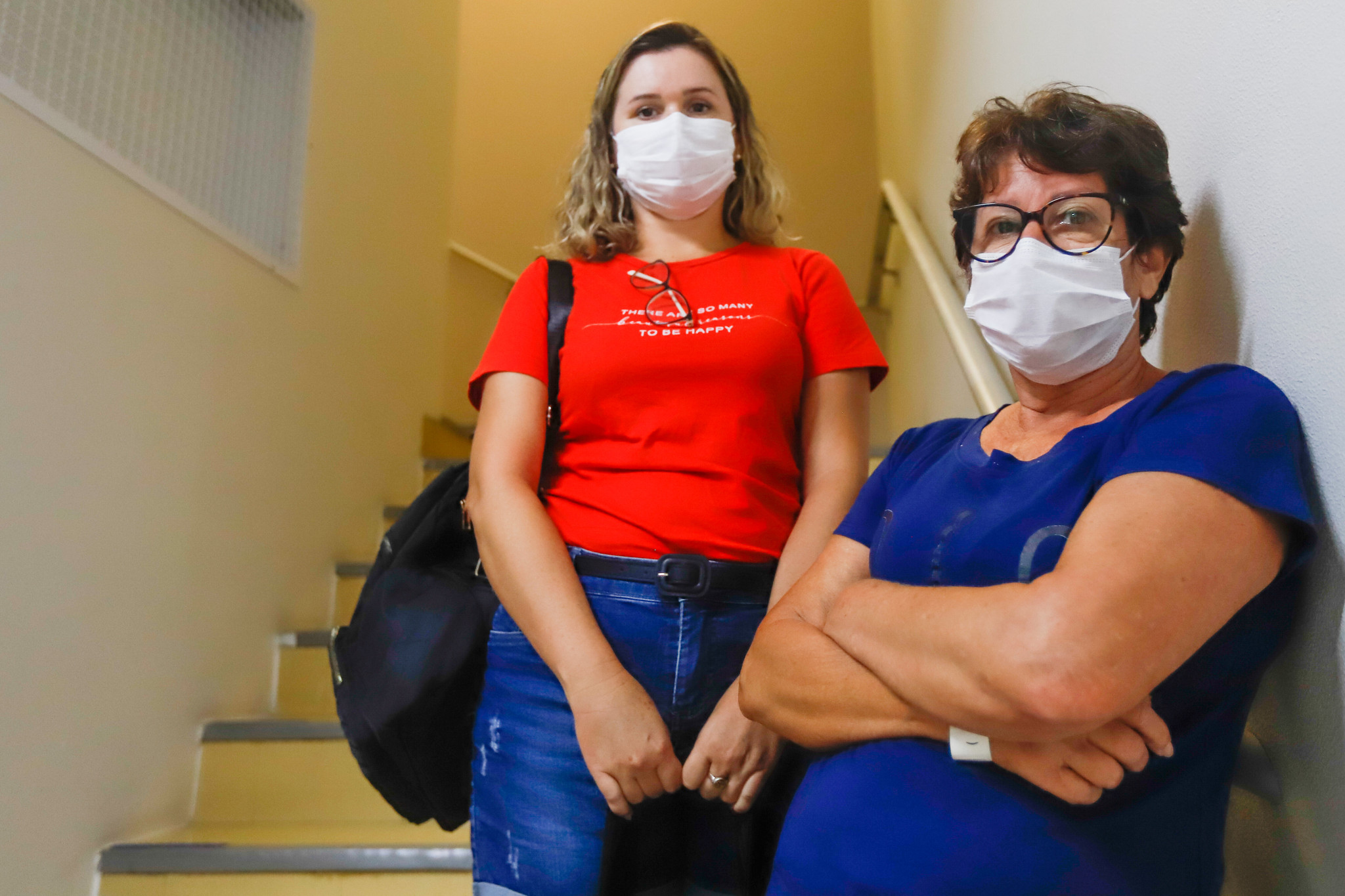 Duas mulheres usando máscaras de proteção posam em uma escada de ambiente interno. A mulher à esquerda veste camiseta vermelha com a frase 'There are so many reasons to be happy', calça jeans e mochila nas costas. A mulher à direita, de braços cruzados, usa óculos e camiseta azul. Ambas olham para a câmera, em uma expressão séria e atenta.
