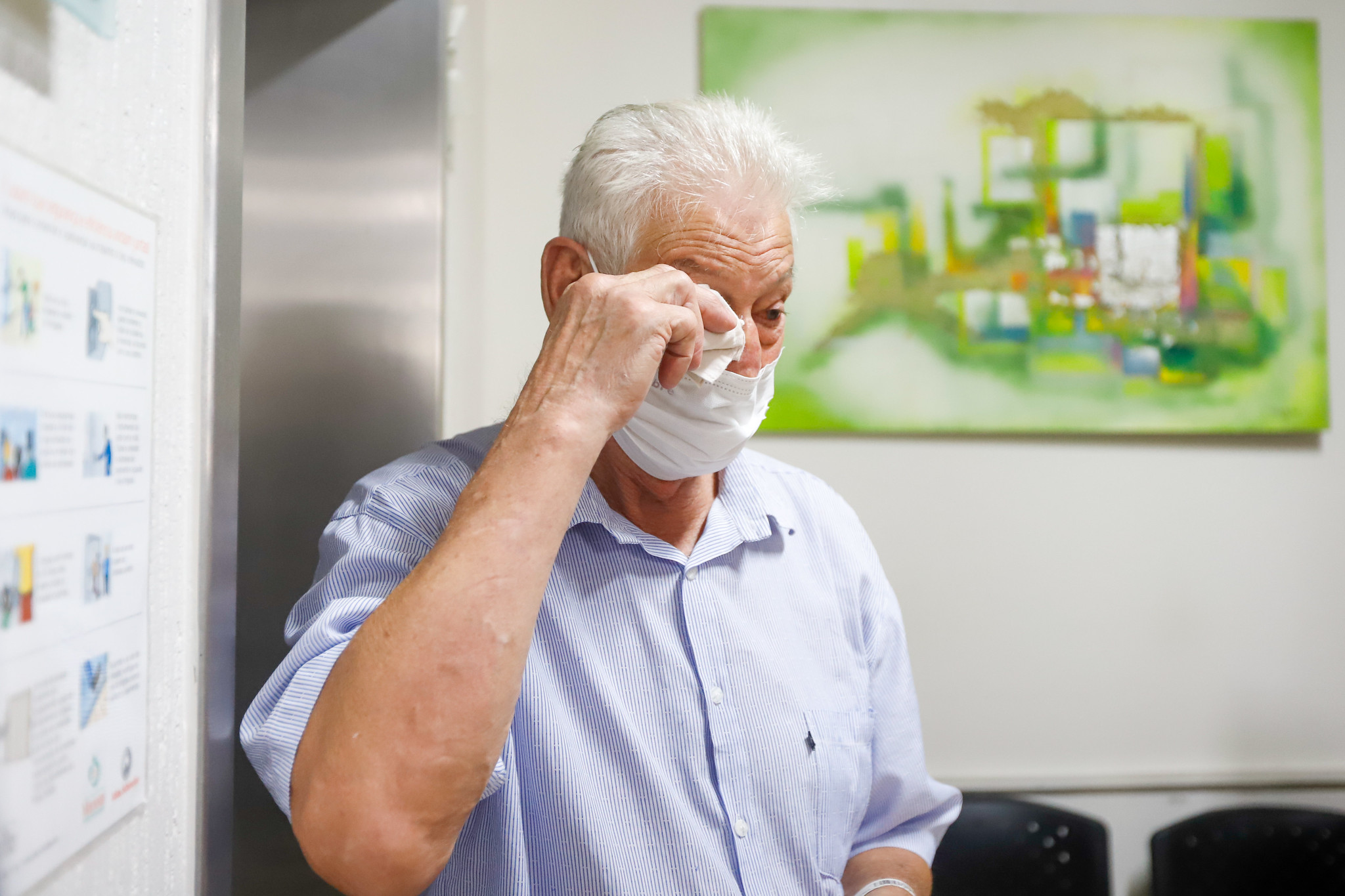 Homem idoso, de cabelos brancos, usando camisa azul clara e máscara de proteção facial branca, enxuga os olhos com um lenço de papel. Ele aparenta estar emocionado. Ao fundo, há uma pintura abstrata em tons de verde e um painel informativo parcialmente visível na parede. A cena transmite um momento íntimo e sensível, possivelmente em ambiente hospitalar ou de saúde.