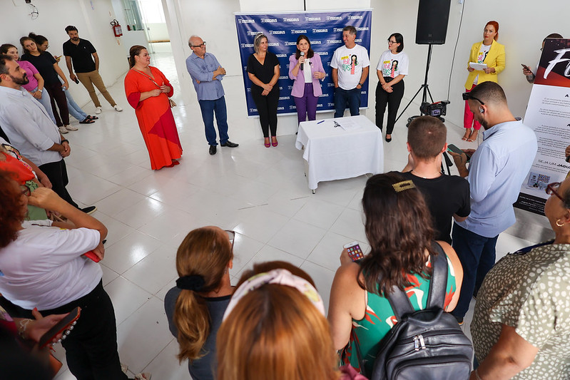 Evento em local fechado com grande participação de pessoas. Uma mulher de blazer lilás fala ao microfone em frente a um painel institucional da Fundação Catarinense de Cultura (FCC), acompanhada por outras autoridades. O público está atento à apresentação, com algumas pessoas fotografando e filmando. O ambiente tem piso branco e iluminação natural, com clima informal e acolhedor.