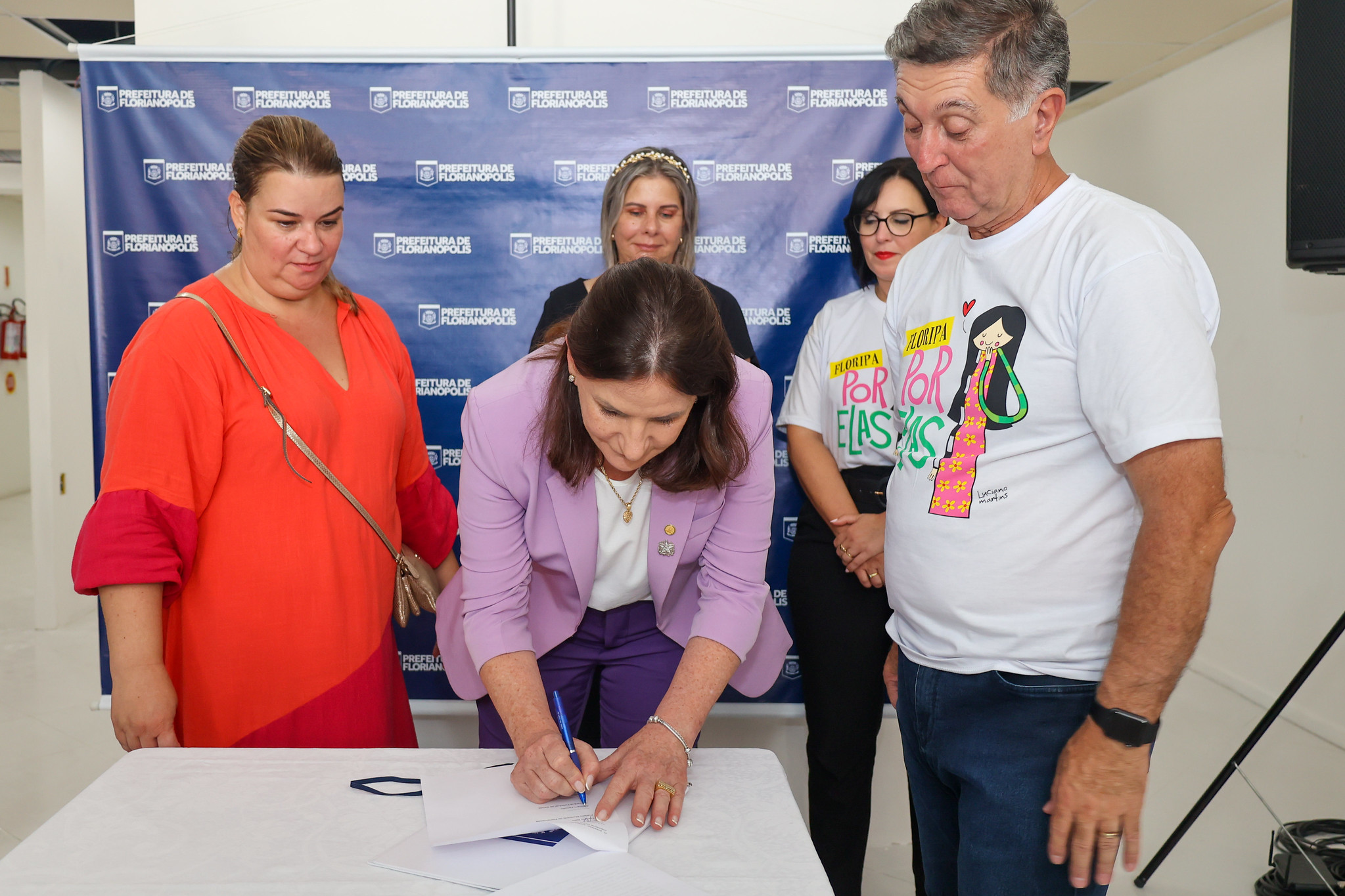 Mulher de blazer lilás e calça roxa assina documento sobre uma mesa branca, cercada por outras pessoas. Ao fundo, um painel azul com o logotipo da Prefeitura de Florianópolis. Duas pessoas vestem camisetas com a frase “Floripa por Elas”. O momento parece marcar uma cerimônia oficial ou assinatura de parceria voltada às mulheres.