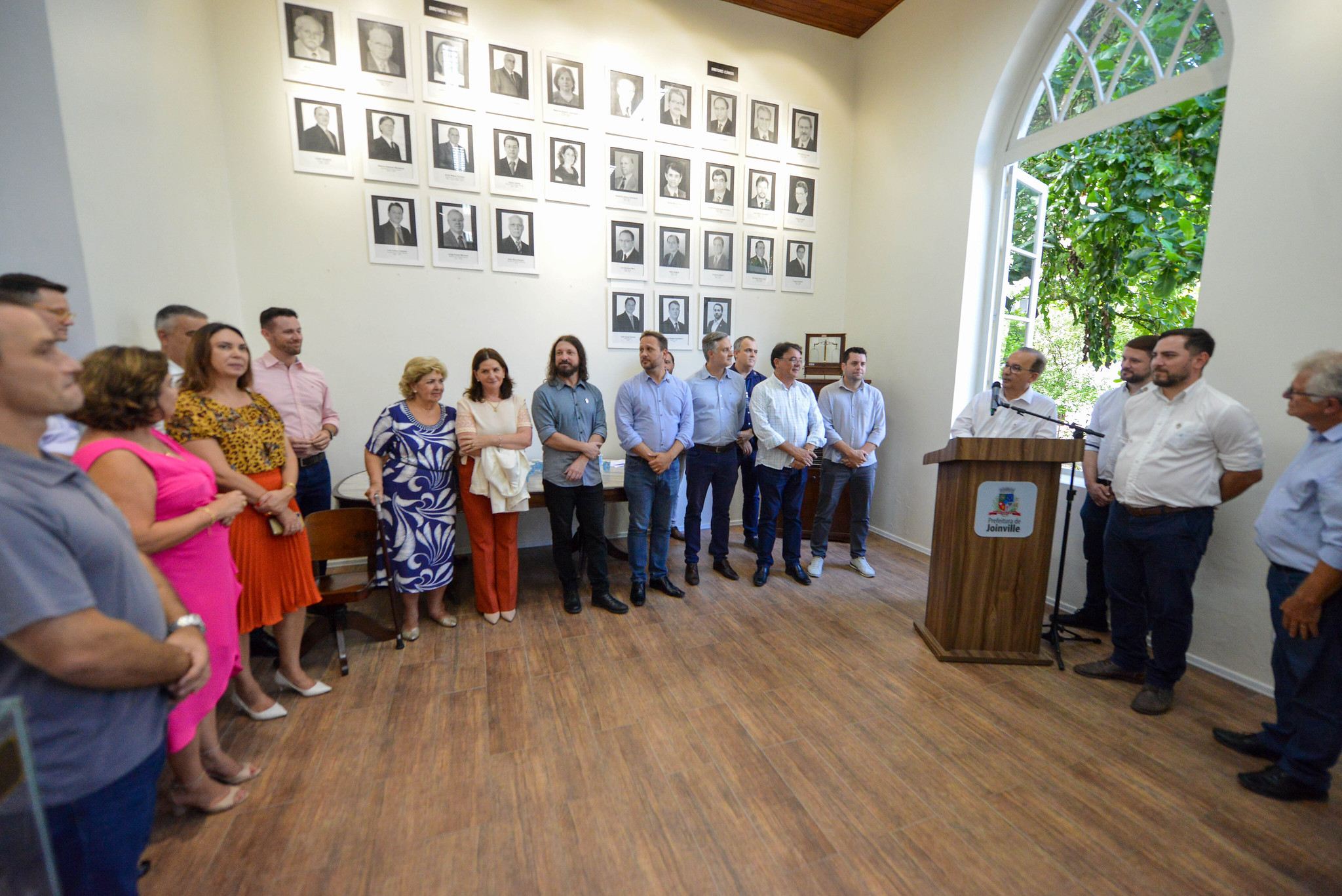 Evento oficial em sala com piso de madeira e paredes decoradas com retratos de ex-prefeitos de Joinville. Um grupo de aproximadamente vinte pessoas está reunido, com parte dos participantes ouvindo um homem falando ao microfone em púlpito com brasão da cidade. A janela ao fundo revela vegetação externa.