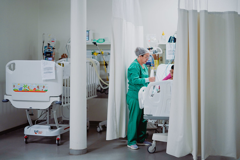 Profissional de saúde em uniforme verde presta atendimento a paciente em leito hospitalar cercado por cortinas, em uma enfermaria equipada com monitores, bombas de infusão e camas hospitalares pediátricas.