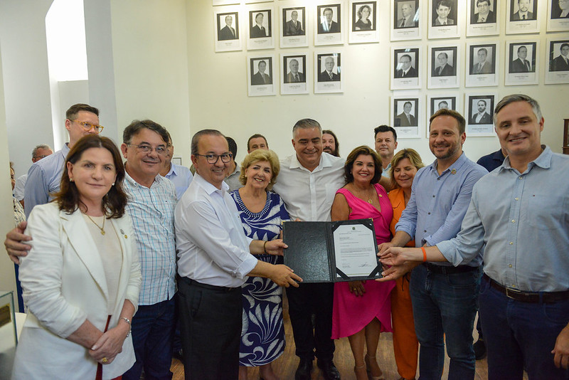 Grupo de pessoas sorridentes posa para foto em uma sala com retratos de autoridades nas paredes. No centro, algumas delas seguram um documento aberto, em clima de celebração. A maioria veste roupas sociais e coloridas, indicando um evento formal ou institucional.