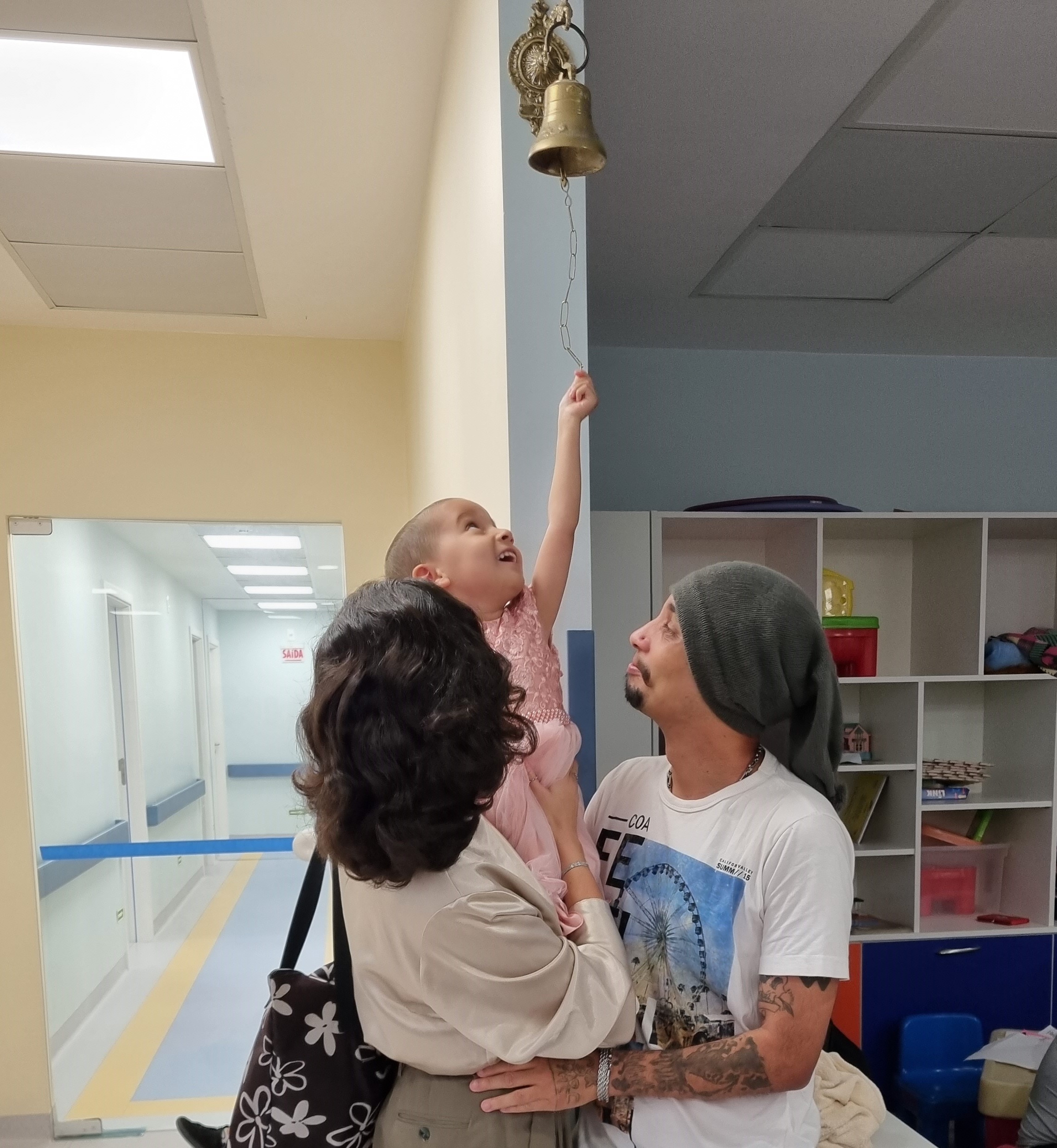 A imagem mostra um momento emocionante em um ambiente hospitalar. Uma criança, no colo de um homem e acompanhada por uma mulher, toca um sino dourado pendurado no alto da parede. A criança olha para cima com expressão de alegria e realização, enquanto o homem e a mulher a observam com carinho e orgulho.  O sino é tradicionalmente usado em hospitais oncológicos para simbolizar o fim do tratamento ou uma conquista importante na jornada contra o câncer. Ao fundo, há um corredor iluminado e, à direita, prateleiras com brinquedos e objetos coloridos, sugerindo que o local pode ser uma ala pediátrica.