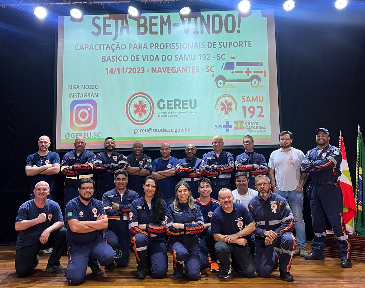 A imagem mostra um grupo de profissionais do SAMU 192 – SC posando juntos no palco, diante de um telão que exibe a mensagem de boas-vindas para a "Capacitação para Profissionais de Suporte Básico de Vida", realizada em 14/11/2023, em Navegantes-SC. No telão também aparecem o logotipo do GEREU (Gerência de Educação em Urgências de Santa Catarina), o endereço de e-mail institucional, o perfil do Instagram e os logotipos do SUS e da Secretaria de Estado da Saúde de Santa Catarina. Os participantes usam uniformes do SAMU e demonstram espírito de equipe, com alguns posando com os braços cruzados e outros agachados na frente. Ao lado do grupo, estão as bandeiras do Brasil e de Santa Catarina.