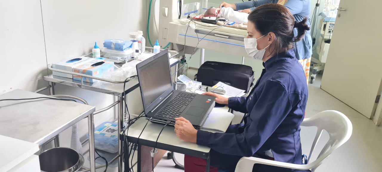 Profissional da saúde usando jaleco azul e máscara facial trabalha em um notebook dentro de uma unidade neonatal. Ao fundo, um bebê recém-nascido está deitado em uma incubadora, sendo cuidado por outra pessoa. A sala tem equipamentos médicos, materiais de enfermagem e iluminação natural vinda da janela.