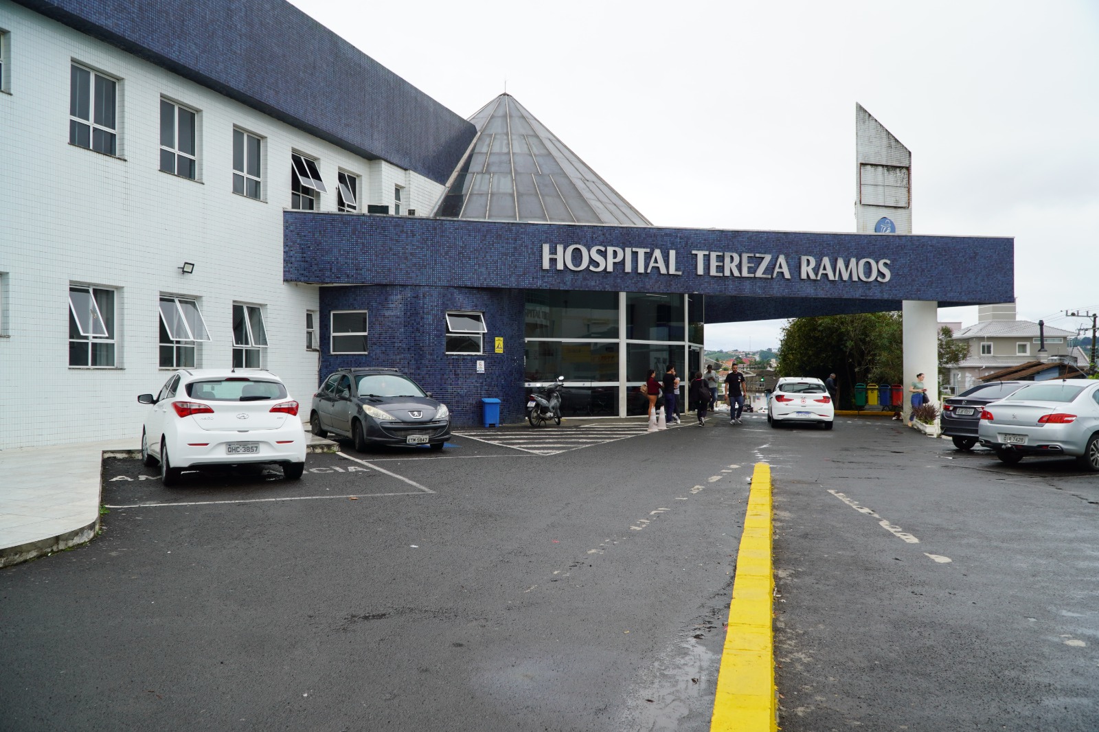 A imagem mostra a entrada principal do Hospital Tereza Ramos, um prédio de fachada com revestimento em pastilhas azul-escuras na parte superior e brancas na parte inferior. No centro, há uma estrutura de vidro e metal em formato piramidal sobre a recepção. Em destaque, a marquise azul com o nome do hospital escrito em letras grandes e brancas.  Na frente, há uma área de estacionamento com carros e uma motocicleta, além de uma faixa amarela que separa a via de acesso. Algumas pessoas estão próximas à entrada, indicando movimento típico de um ambiente hospitalar. Ao fundo, é possível ver parte do bairro, com casas e postes, sob um céu nublado.