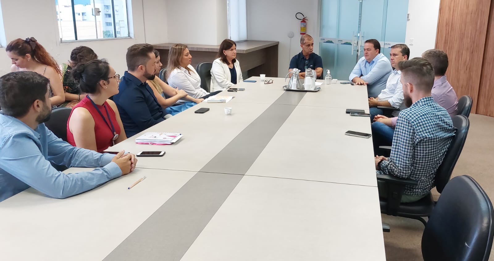 Reunião formal com várias pessoas sentadas ao redor de uma mesa retangular em uma sala de reuniões corporativa. Participantes de diferentes idades e gêneros estão atentos à conversa, com celulares, cadernos e materiais de trabalho à frente. Ao fundo, há janelas, uma porta de vidro e uma bandeja com garrafas de café e copos.