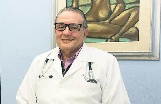 Homem usando óculos e jaleco branco com estetoscópio no pescoço, sorrindo em consultório médico, com quadro artístico ao fundo.