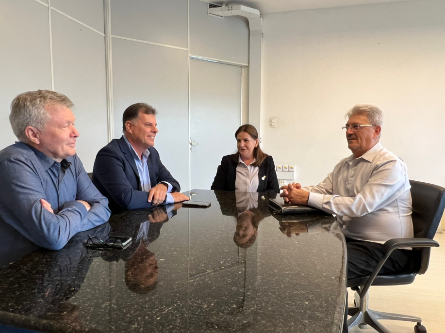Quatro pessoas estão reunidas em torno de uma mesa de granito preta em uma sala de reuniões. À esquerda da imagem, estão dois homens de terno azul escuro. À direita, uma mulher de blazer preto e camisa branca está sentada ao lado de outro homem de camisa branca. Todos estão sorrindo e em conversa, com expressão amigável. Há itens sobre a mesa, como óculos, celular e uma pasta.