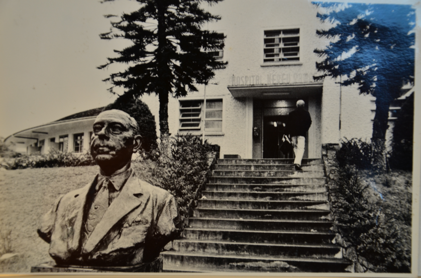 Fotografia em preto e branco de um hospital, com escadaria central e fachada onde se lê “Hospital Nossa Senhora”. Em destaque no primeiro plano, há um busto de um homem idoso de terno e gravata, provavelmente homenageado pela instituição. Um homem caminha escada acima em direção à entrada principal. A cena é rodeada por árvores altas e vegetação.