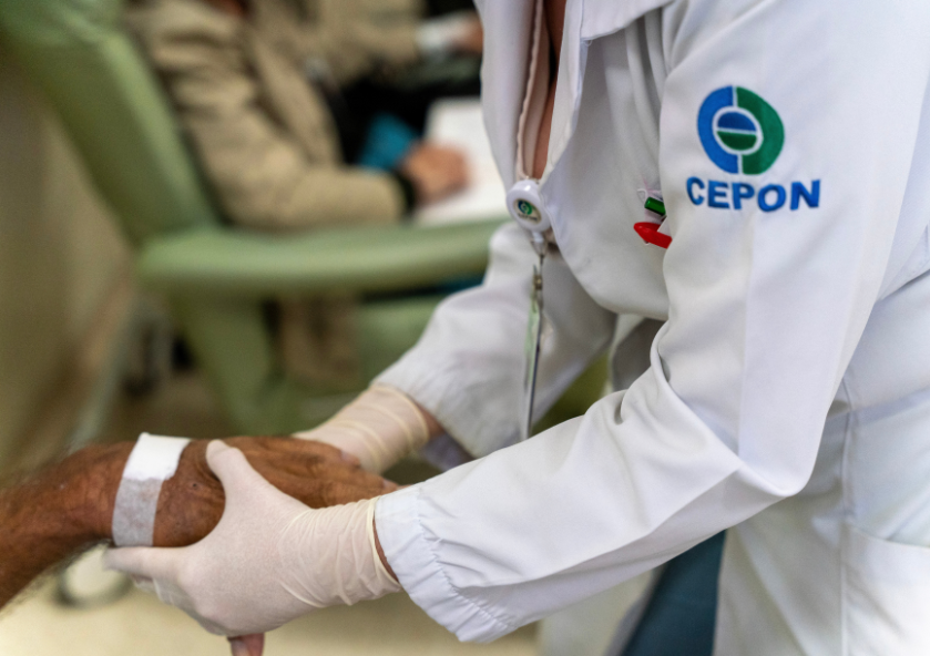 Profissional da saúde do CEPON, com jaleco branco e luvas, segura cuidadosamente a mão de um paciente com curativo no braço, em ambiente clínico, demonstrando cuidado e atenção humanizada.
