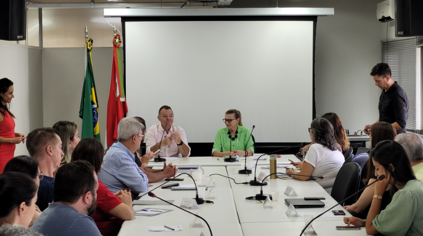 Reunião formal realizada em uma sala de conferência. Cerca de 20 pessoas estão sentadas ao redor de uma grande mesa branca, com microfones distribuídos. Ao fundo, um homem e uma mulher, que parecem conduzir a reunião, estão sentados ao centro, próximos a um projetor e a duas bandeiras: do Brasil e de Santa Catarina. Algumas pessoas fazem anotações, outras usam celulares ou prestam atenção aos falantes. Há uma tela branca grande ao fundo, indicando a possibilidade de uma apresentação. O ambiente é bem iluminado, com janelas e persianas ao lado direito.