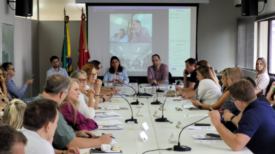 Reunião realizada em uma sala com cerca de 20 pessoas sentadas ao redor de uma mesa retangular branca, equipada com microfones. No fundo da sala, há uma projeção de uma videoconferência com outros participantes em chamada. Bandeiras do Brasil e de Santa Catarina estão posicionadas no canto. Os participantes analisam documentos, fazem anotações e alguns utilizam celulares, demonstrando concentração na discussão.