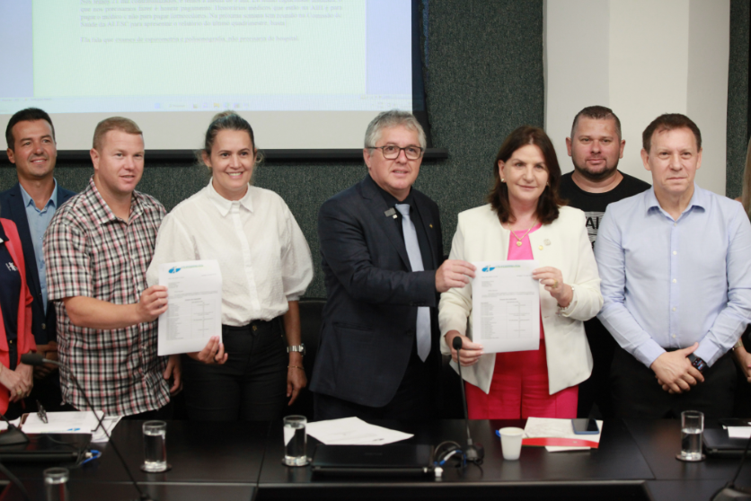 Grupo de pessoas em uma sala de reuniões posando para foto, com duas delas segurando documentos oficiais. Ao fundo, tela de projeção com texto. Clima de formalidade e celebração de parceria ou assinatura de acordo.