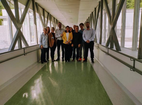 Grupo de pessoas posando para foto em um corredor envidraçado de uma unidade de saúde. O local é iluminado por luz natural e possui piso verde e corrimãos nas laterais. Ao centro, uma mulher com blazer amarelo e outras pessoas com trajes sociais.