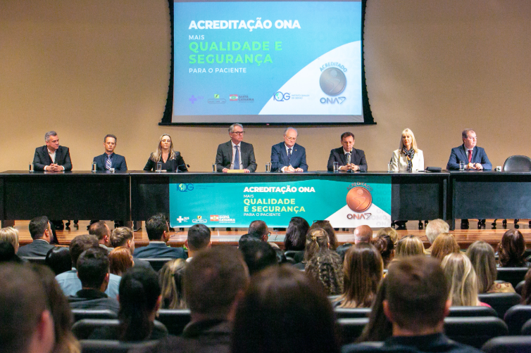 Autoridades sentadas à mesa principal durante cerimônia de acreditação ONA 3, com público presente em auditório. Ao fundo, telão com os dizeres: 'Acreditação ONA – Mais qualidade e segurança para o paciente'.