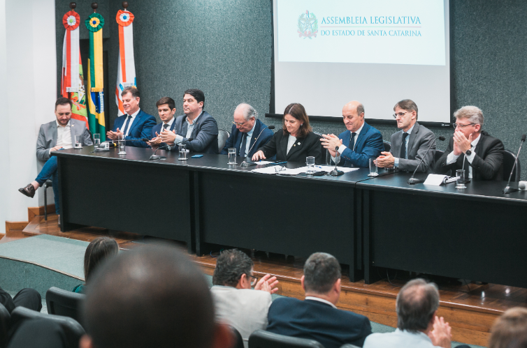 Autoridades reunidas em mesa oficial durante evento na Assembleia Legislativa do Estado de Santa Catarina, com bandeiras ao fundo e público assistindo.