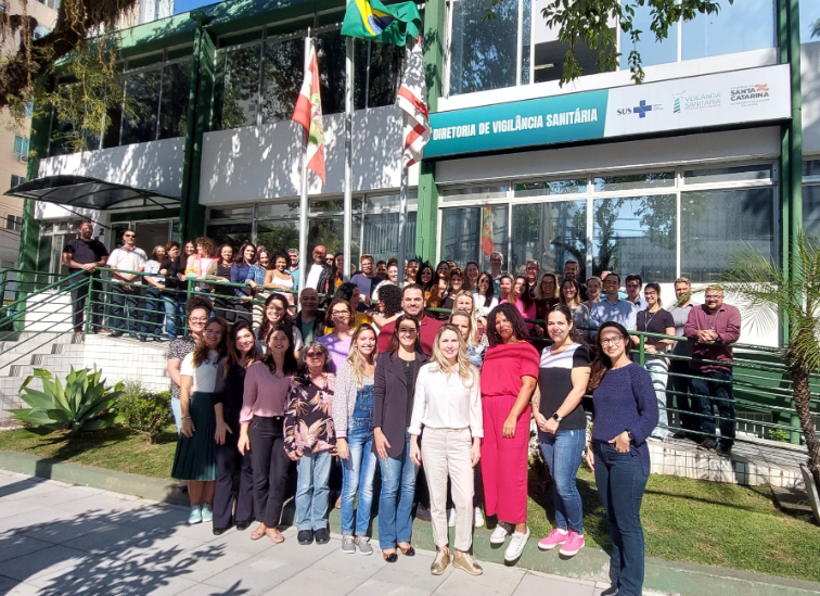 Grande grupo de profissionais posa para foto em frente à sede da Diretoria de Vigilância Sanitária de Santa Catarina, em um dia ensolarado. Três bandeiras estão hasteadas e há placas do SUS e do Governo do Estado ao fundo.