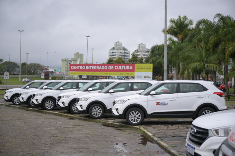 Fila de veículos brancos oficiais do Governo de Santa Catarina estacionados em frente ao Centro Integrado de Cultura, em Florianópolis. Ao fundo, uma placa vermelha indica as atividades do local, como museus, cinema, teatro e biblioteca. Palavras "Santa Catarina" e o brasão do governo estão estampados nas portas dos carros.
