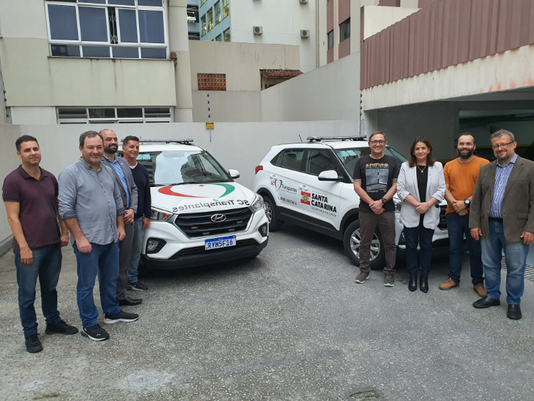 Grupo de nove pessoas posa em frente a dois carros brancos com a identidade visual do SC Transplantes e da Secretaria da Saúde de Santa Catarina. Os veículos estão estacionados em uma área externa entre prédios, simbolizando apoio logístico às ações de transplante no estado.