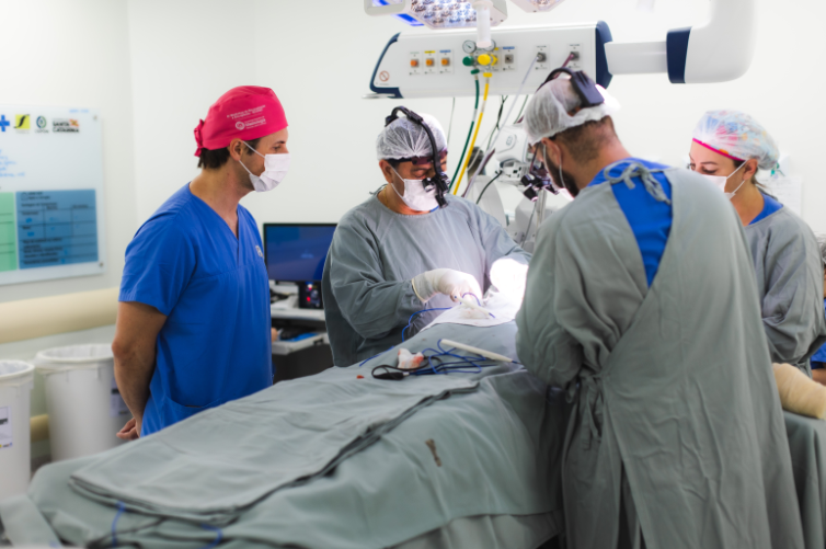 Profissionais de saúde usando roupas cirúrgicas, máscaras e toucas realizando procedimento em centro cirúrgico, com foco de luz sobre o paciente.