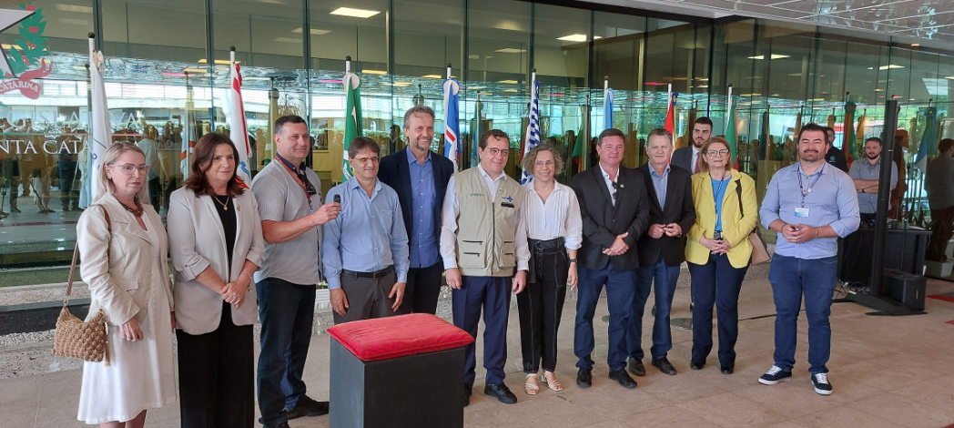 Grupo de autoridades e representantes posando para foto em frente à entrada da Assembleia Legislativa de Santa Catarina, com bandeiras ao fundo e um pedestal com toalha vermelha à frente.