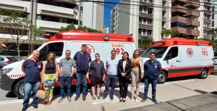 A imagem mostra um grupo de nove pessoas posando em frente a duas ambulâncias do SAMU (Serviço de Atendimento Móvel de Urgência), ambas identificadas com o número de emergência 192 e a inscrição “Unidade de Suporte Básico”. As ambulâncias têm a pintura branca com detalhes em vermelho e o logotipo do SAMU, além da indicação de “Santa Catarina”. O grupo é formado por homens e mulheres, alguns vestindo uniformes do SAMU e outros em trajes casuais ou formais. O cenário de fundo inclui prédios residenciais, árvores e carros estacionados, sugerindo que a foto foi tirada em uma área urbana em um dia ensolarado.