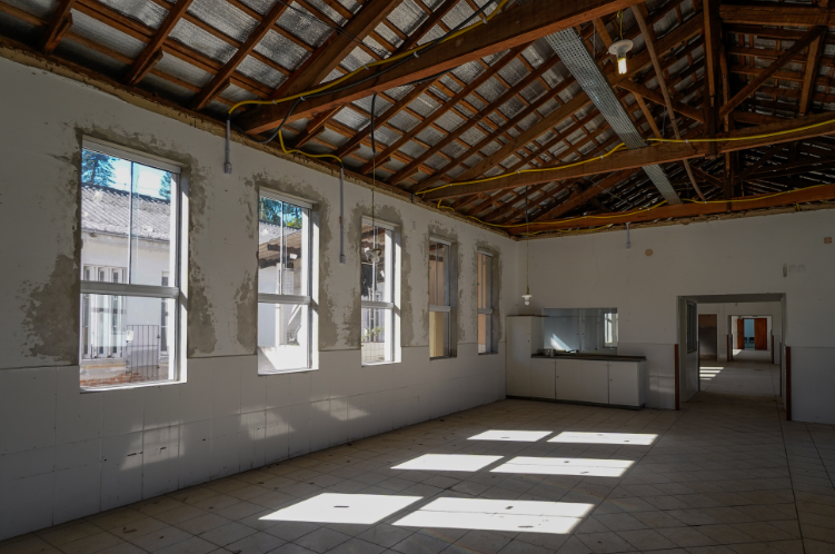 A imagem mostra um salão amplo e iluminado por várias janelas altas em uma das paredes laterais. As paredes apresentam sinais de desgaste, com partes do reboco descascadas ao redor das janelas. O teto é de estrutura de madeira exposta, com telhas cerâmicas e algumas instalações elétricas visíveis, incluindo fiação amarela e lâmpadas penduradas. O piso é de cerâmica clara, disposto em padrão diagonal, e há um balcão com armários brancos e pia ao fundo, próximo a uma abertura que conecta a outro espaço. Ao fundo, por um corredor, é possível ver outra área iluminada. O ambiente aparenta estar em fase de reforma ou necessitando de manutenção.