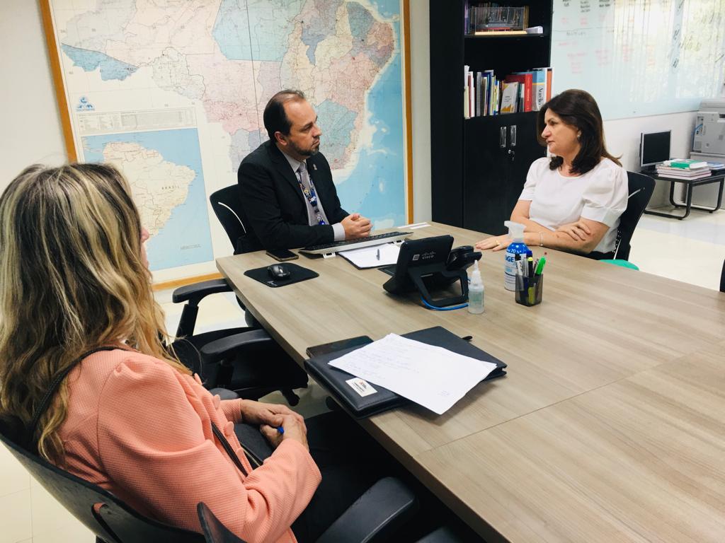 Três pessoas sentadas ao redor de uma mesa retangular em uma reunião. Ao fundo, há um grande mapa do Brasil na parede. Uma mulher de blusa branca, um homem de terno e uma mulher de blazer coral estão engajados em conversa, com anotações, telefone e itens de escritório sobre a mesa.