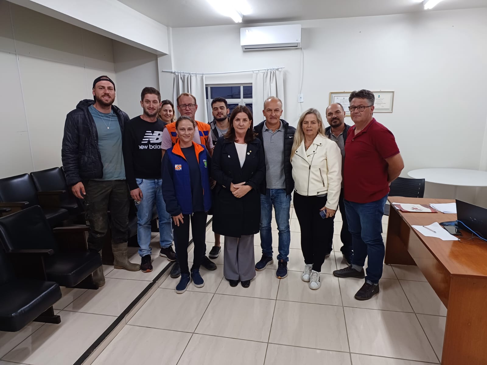 Grupo de homens e mulheres posando para foto em sala de reuniões, com alguns usando uniformes da Defesa Civil, durante visita a cidades afetadas no Alto Vale.