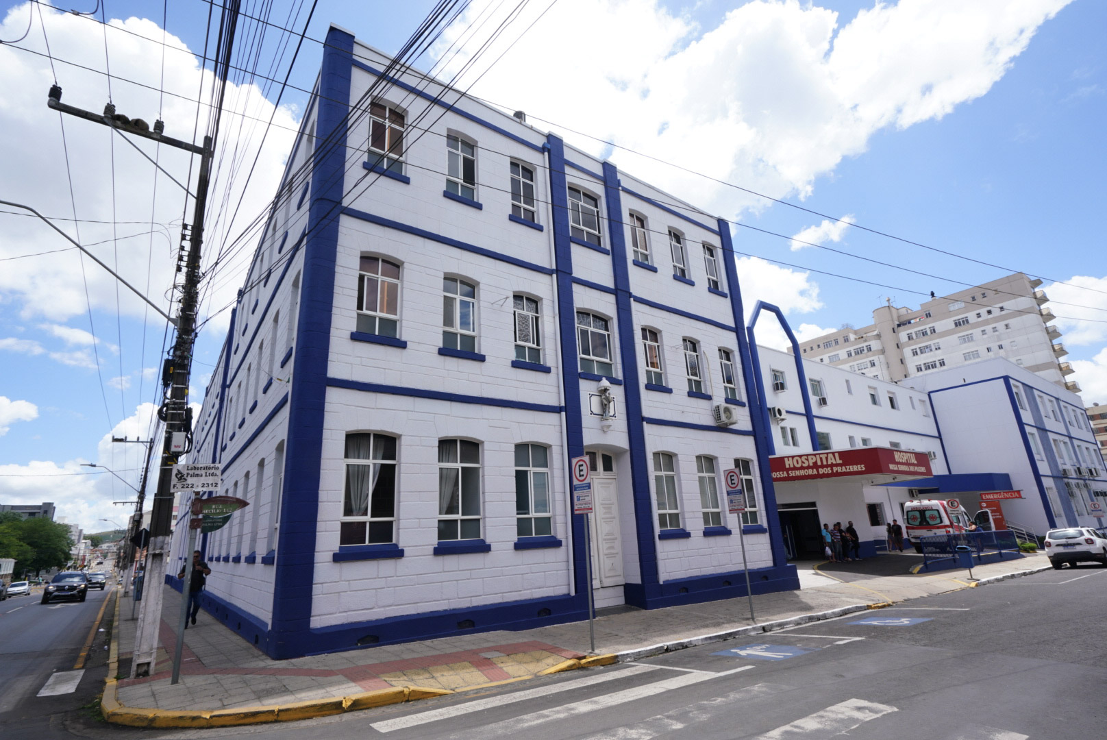 Prédio de três andares pintado de branco com detalhes em azul, localizado em esquina urbana. À direita, há uma entrada com letreiro vermelho onde se lê “HOSPITAL NOSSA SENHORA DOS PRAZERES” e “EMERGÊNCIA”, com uma ambulância estacionada na entrada e algumas pessoas próximas. O céu está claro com nuvens, e postes com muitos fios elétricos cruzam a imagem. Há também placas de trânsito e uma faixa de pedestres em frente ao hospital.
