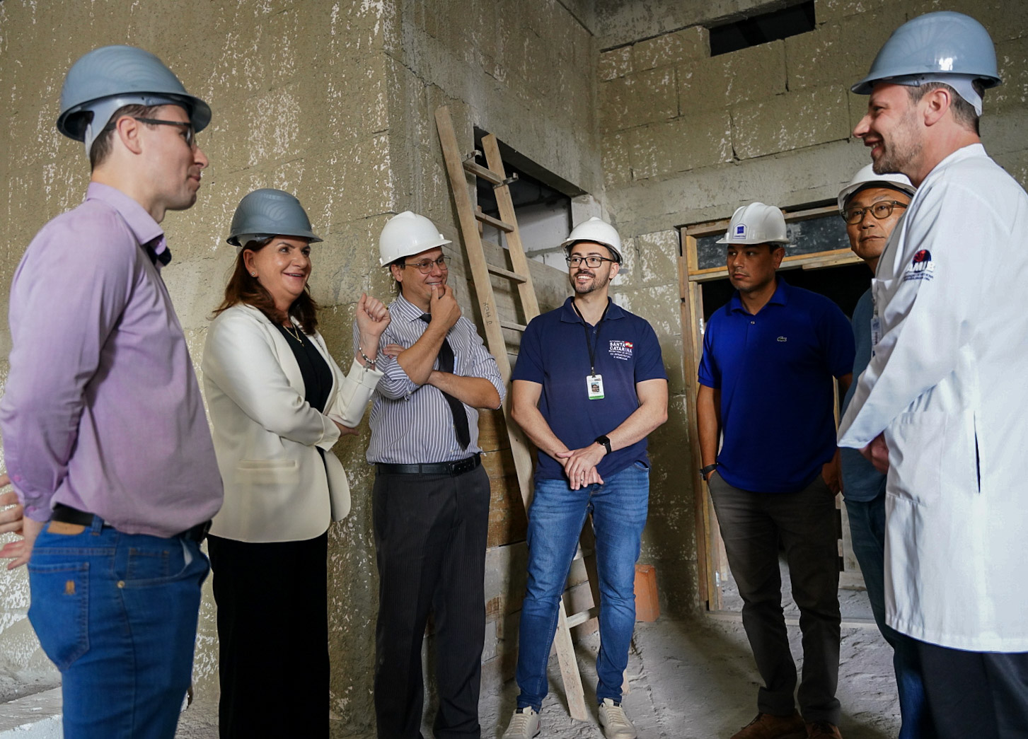 Grupo de sete pessoas, todos usando capacetes de segurança, está reunido em uma obra com paredes de concreto aparente. Entre eles, há uma mulher de blazer branco sorrindo e gesticulando, interagindo com os demais, que são majoritariamente homens. Alguns vestem roupas formais, outros estão com uniforme de trabalho ou jaleco. Ao fundo, vê-se uma escada de madeira encostada na parede. O ambiente sugere uma vistoria ou reunião técnica sobre o andamento da construção.