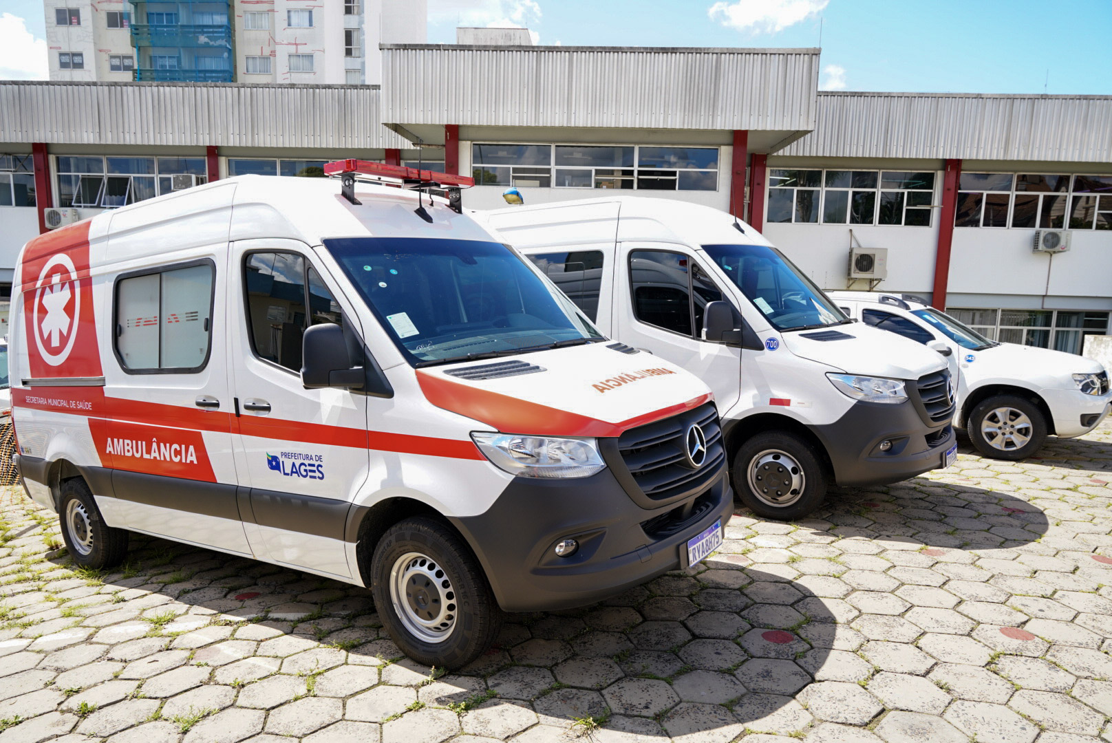 Duas ambulâncias brancas modelo van Mercedes-Benz estão estacionadas lado a lado em calçamento de pedras sextavadas. A da frente possui adesivos vermelhos, logotipo da “Prefeitura de Lages” e inscrição “AMBULÂNCIA” na lateral e capô. Ambas possuem luzes de emergência no teto. Ao fundo, um prédio com fachada branca e detalhes vermelhos, janelas espelhadas e aparelhos de ar-condicionado fixados na parede. Há também outros veículos estacionados ao lado das ambulâncias. O céu está limpo e azul, indicando dia ensolarado.