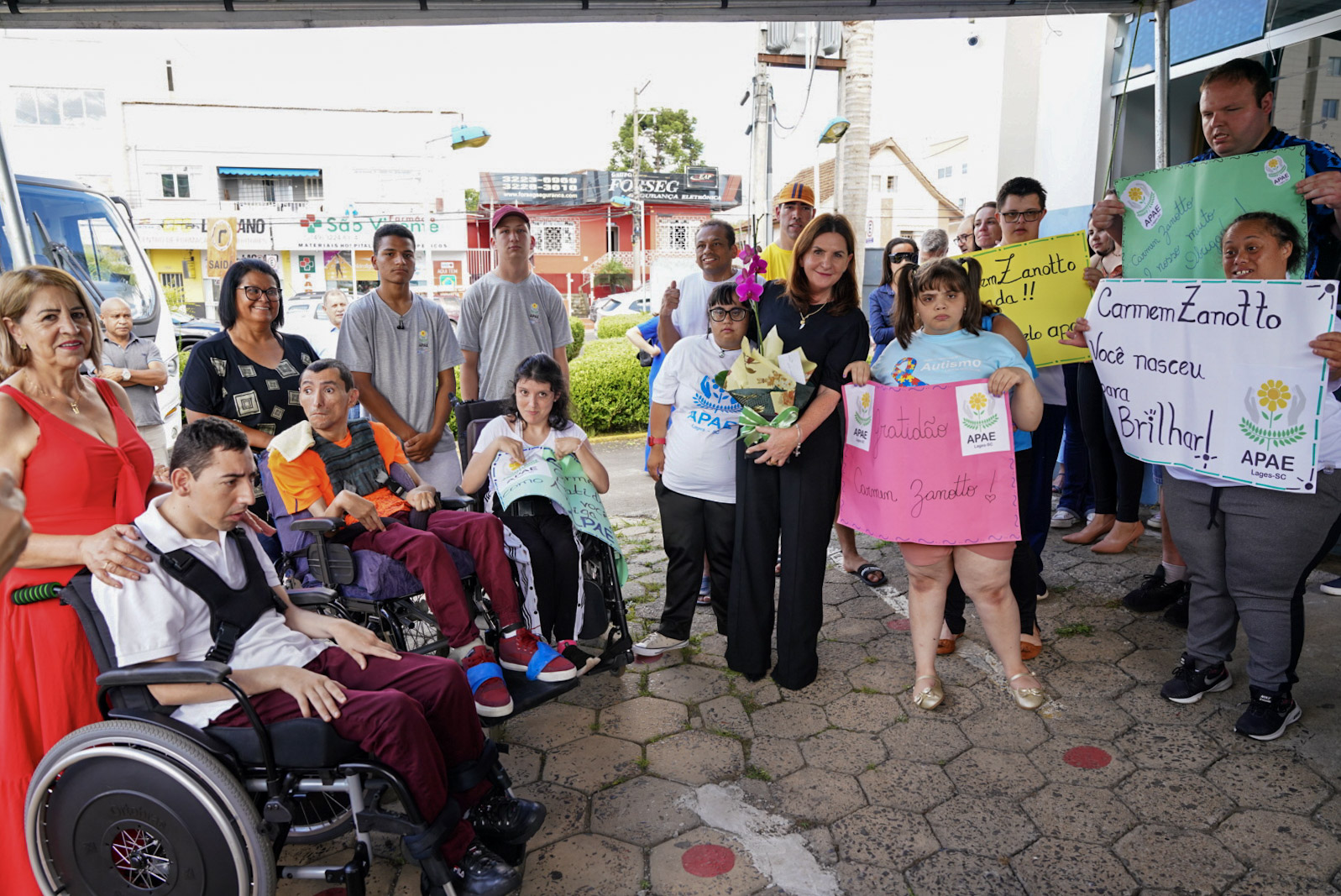 Grupo diverso de pessoas, incluindo alunos da APAE, profissionais e visitantes, está reunido em frente a um prédio, sob cobertura. Alguns dos presentes estão em cadeiras de rodas, outros seguram cartazes coloridos com mensagens de agradecimento à "Carmen Zanotto", como “Você nasceu para brilhar!” e “Gratidão”. Ao centro, uma mulher de roupa preta segura um buquê de flores e sorri. O grupo transmite clima de acolhimento e celebração. Ao fundo, é possível ver uma calçada, veículos e comércios, incluindo uma loja com placa "São José".