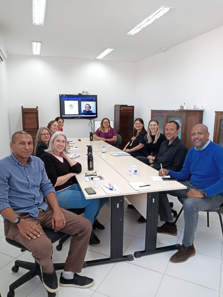 Grupo de pessoas sentadas ao redor de uma mesa de reunião, com cadernos e canecas à frente. Ao fundo, uma televisão transmite videoconferência com participante remoto. O ambiente é iluminado, com armários de madeira encostados na parede.