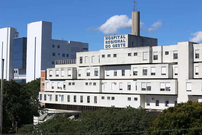 Fachada do Hospital Regional do Oeste em um dia ensolarado, com céu azul e poucas nuvens. O prédio é branco, de múltiplos andares, e possui uma placa visível no topo com o nome da instituição.