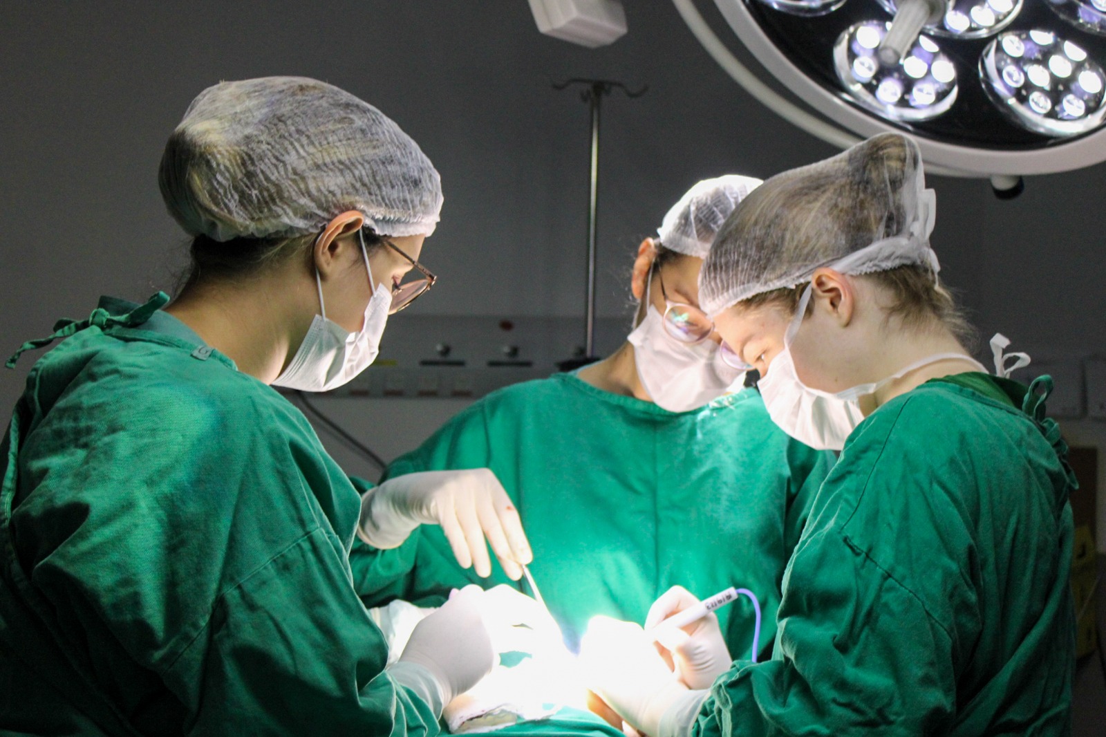 Três profissionais de saúde realizando um procedimento cirúrgico em centro cirúrgico, usando roupas estéreis verdes, toucas e máscaras, sob luz intensa.