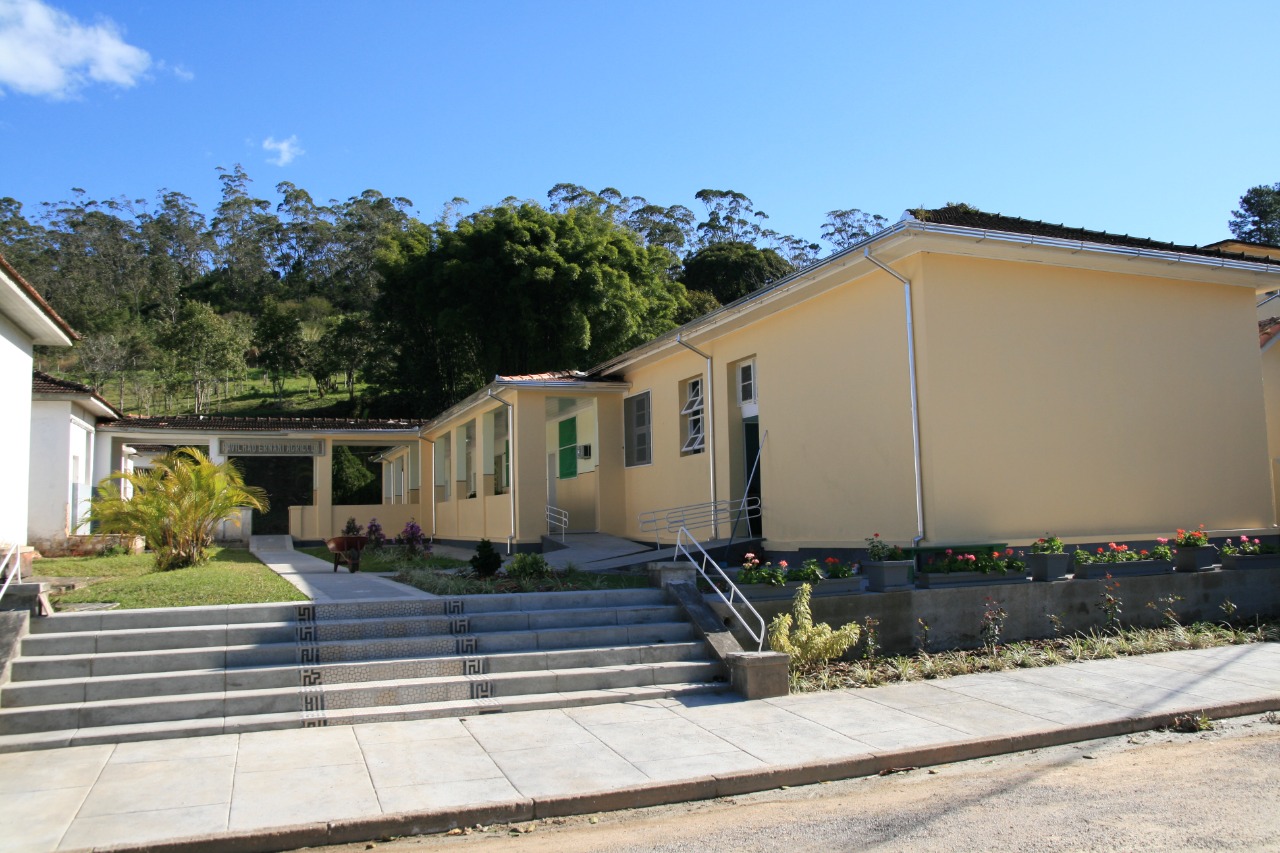 Imagem do Hospital Santa Tereza, com fachada em tom amarelo-claro. O prédio é térreo, com rampas de acesso e escadas na entrada. Há vasos com flores coloridas na frente e um jardim com plantas e grama. Ao fundo, uma área arborizada com muitas árvores e céu azul com poucas nuvens.