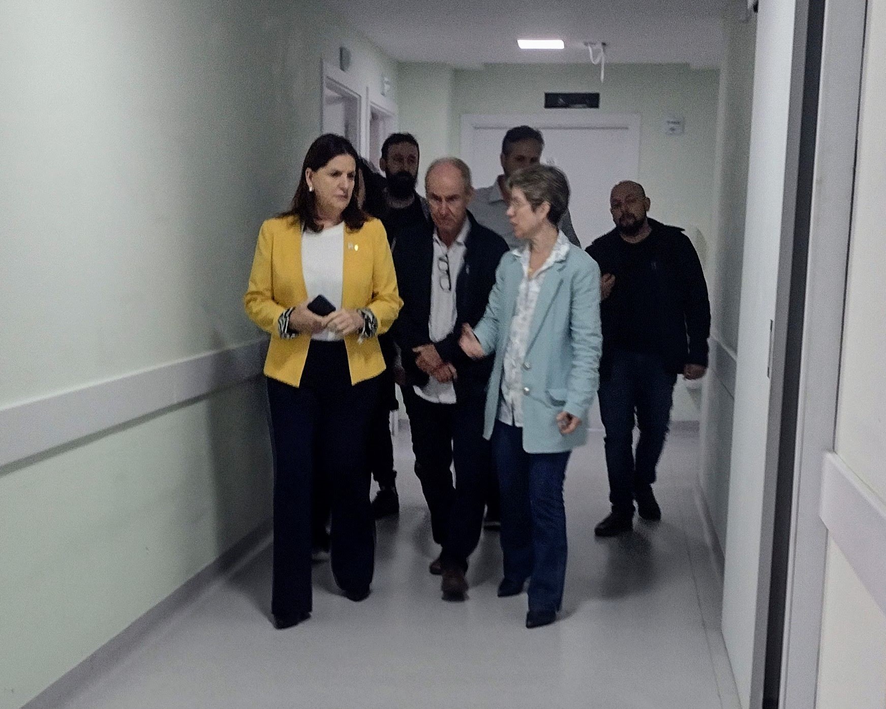 Grupo de pessoas caminha por corredor interno de unidade de saúde. À frente, duas mulheres conversam enquanto os demais acompanham, incluindo homens em trajes sociais e casuais. O ambiente é iluminado e com paredes claras.