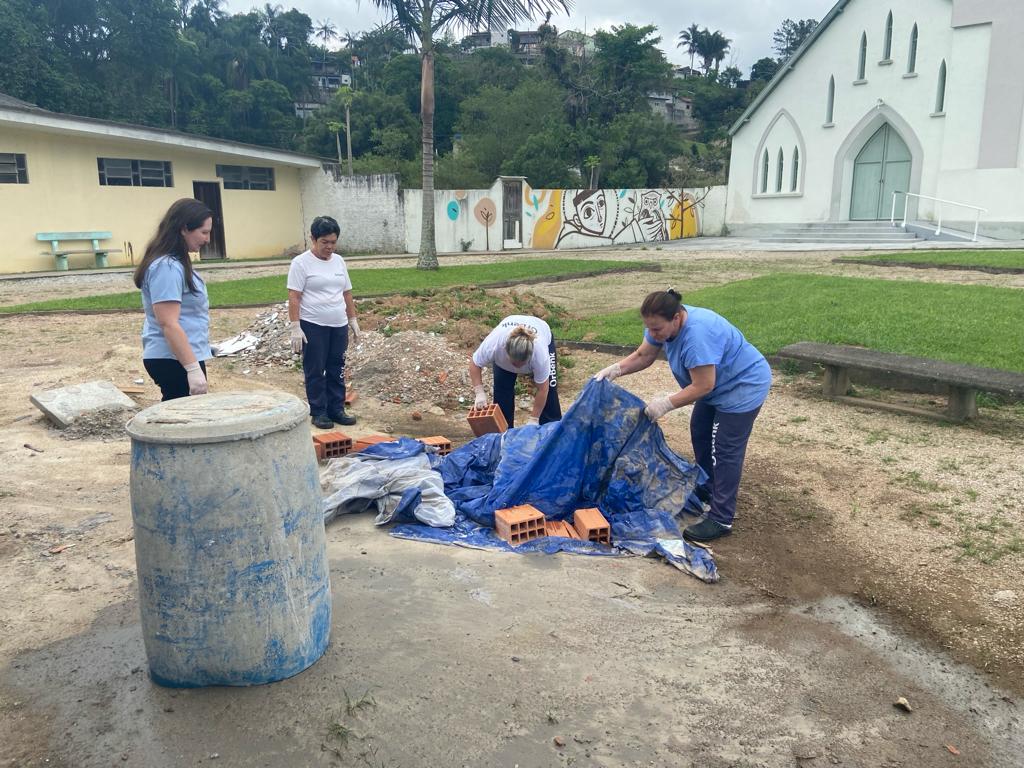 Quatro mulheres realizam trabalho manual em área externa, lidando com tijolos cobertos por uma lona azul. Duas delas seguram a lona, enquanto as outras observam e ajudam. Ao fundo, há um prédio amarelo, um mural colorido, árvores e uma igreja branca com portas verdes.