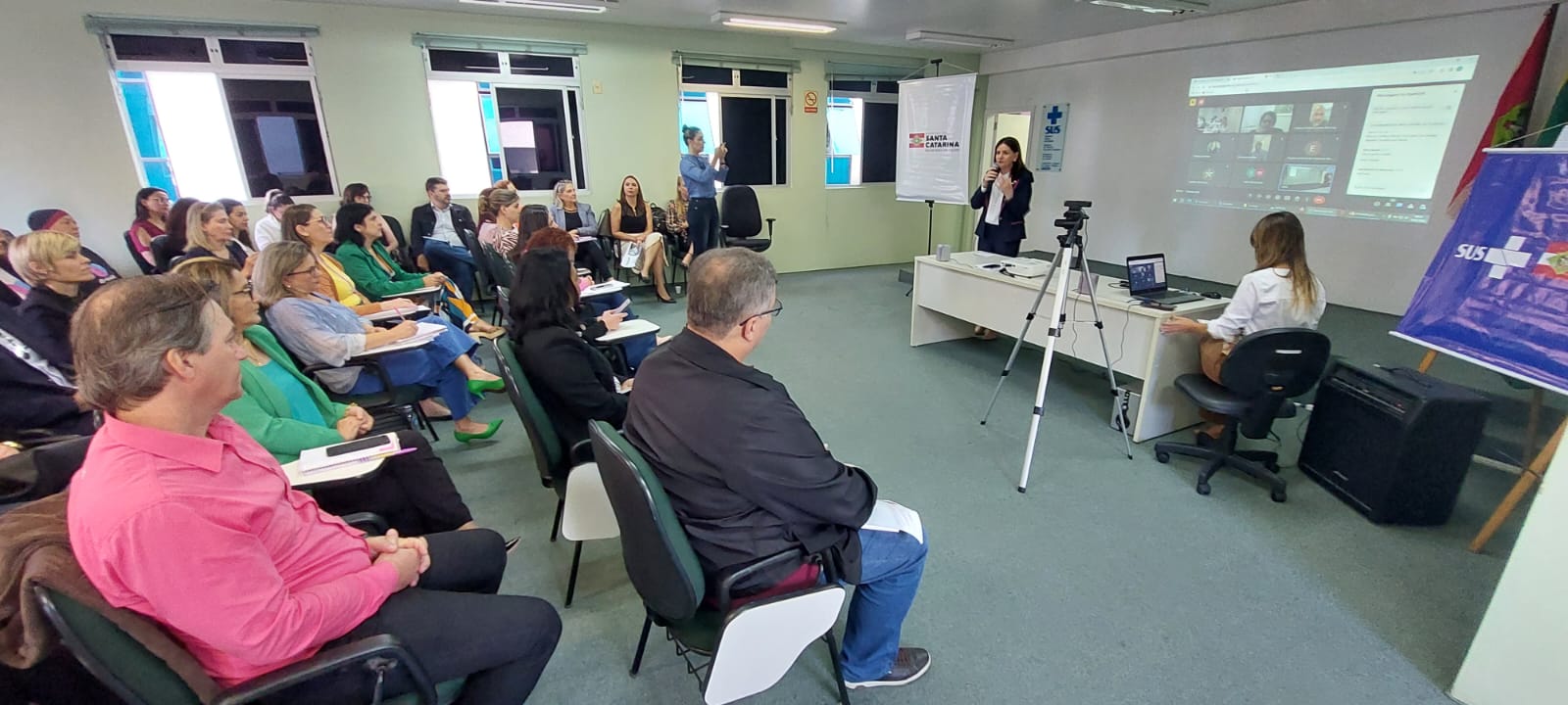 Reunião presencial com várias pessoas sentadas em uma sala, assistindo à apresentação de uma mulher em pé com microfone, ao lado de uma mesa com notebook e tripé de câmera. Uma mulher também está sentada operando o computador. Ao fundo, há uma projeção de uma videoconferência com participantes online e banners do SUS e do Governo de Santa Catarina.