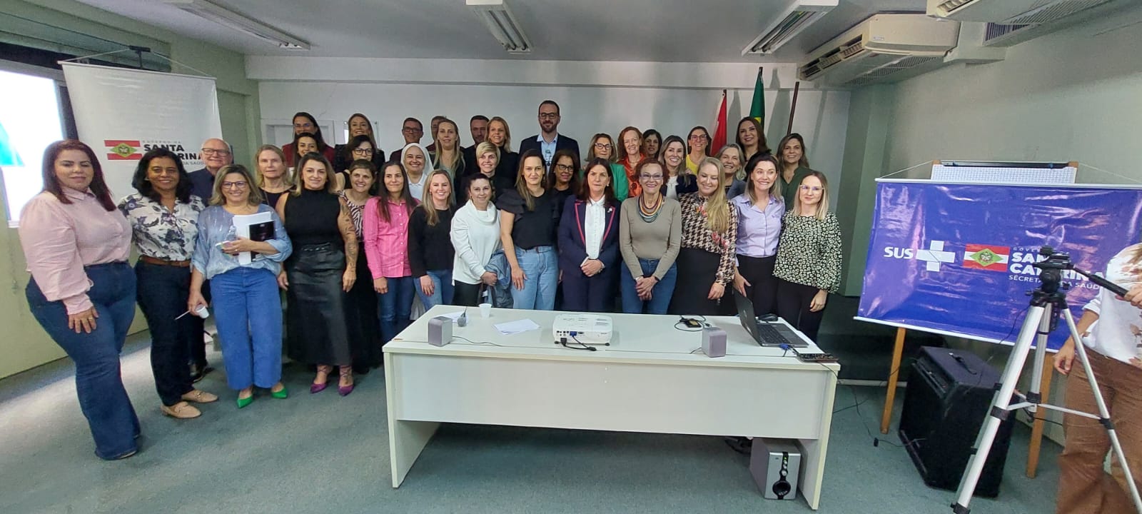 Foto de grupo tirada ao final de um evento em sala de reuniões. Diversas pessoas estão reunidas, sorrindo para a câmera, em pé atrás de uma mesa com projetor, notebook e caixas de som. Ao fundo, há bandeiras e banners do SUS e do Governo de Santa Catarina.