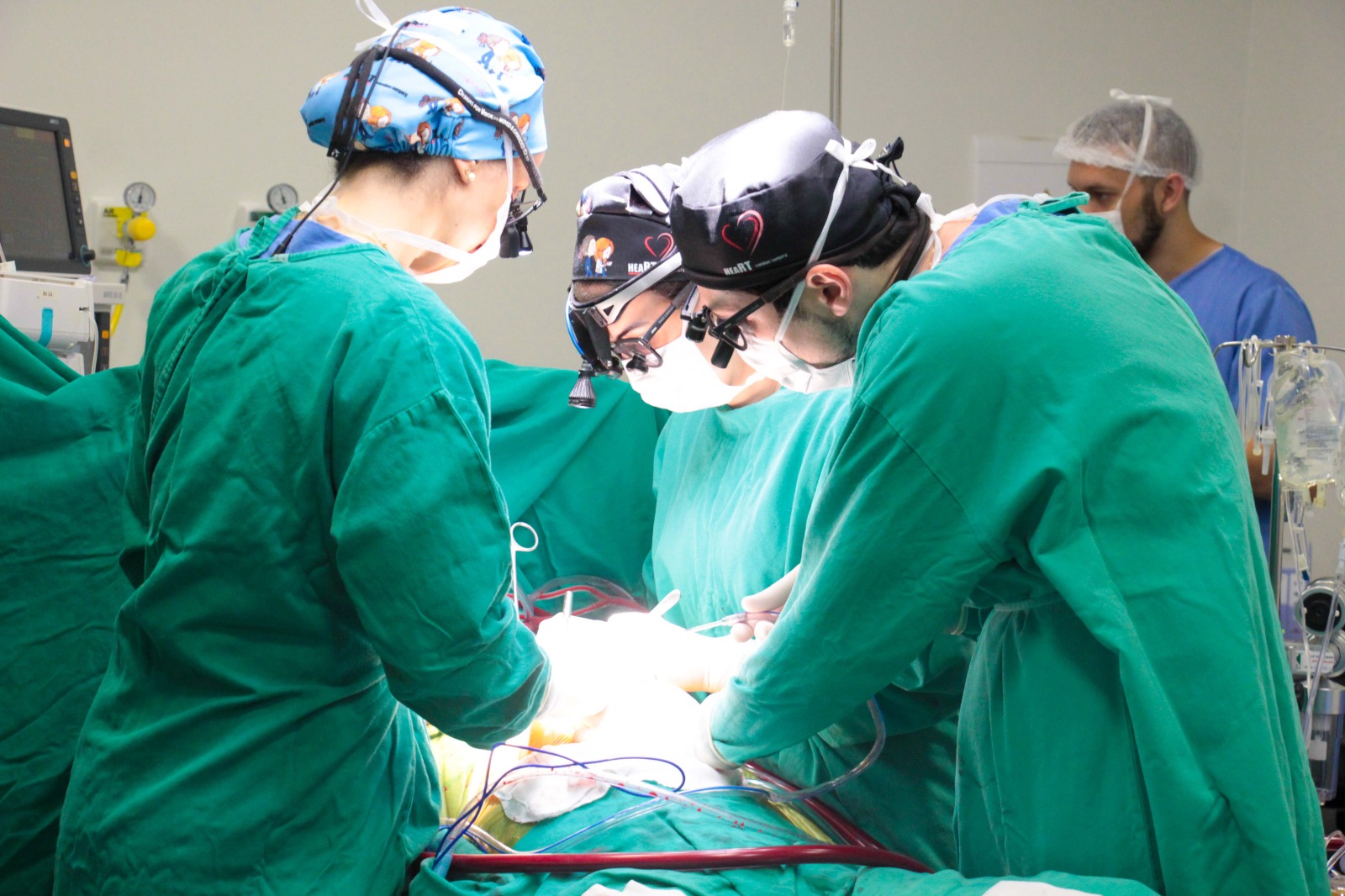 Equipe de cirurgiões vestidos com aventais e toucas verdes realizando procedimento cirúrgico em centro cirúrgico, com luz intensa iluminando a área da operação.
