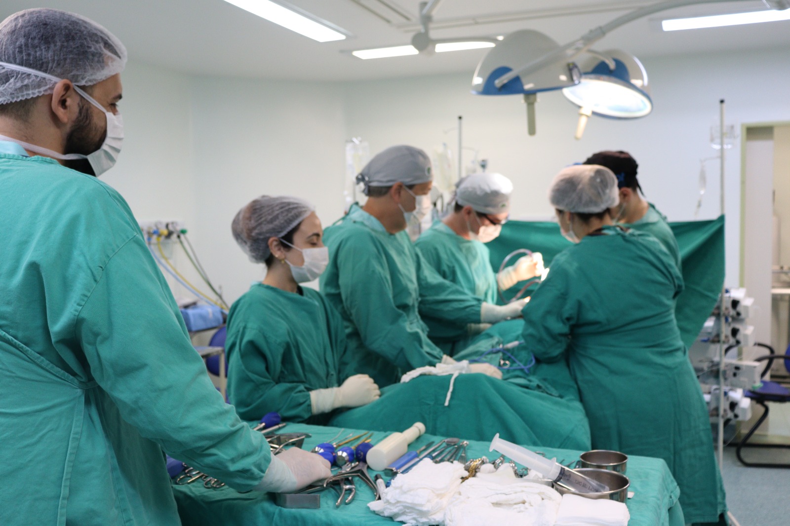 Profissionais de saúde com roupas cirúrgicas realizando procedimento em sala de cirurgia equipada, com instrumentos médicos sobre a mesa.