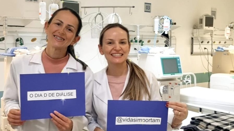 Duas profissionais da saúde vestindo jalecos brancos sorriem segurando cartazes com as frases “O DIA D DE DIÁLISE” e “@vidasimportam”. Ao fundo, há máquinas de hemodiálise e leitos hospitalares, indicando um ambiente clínico especializado.